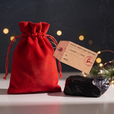 A red velvet drawstring gift pouch with a kraft paper tag reading 'Official North Pole Delivery' sits on a white surface next to a black wrapped item, with warm blurred lights in the background.